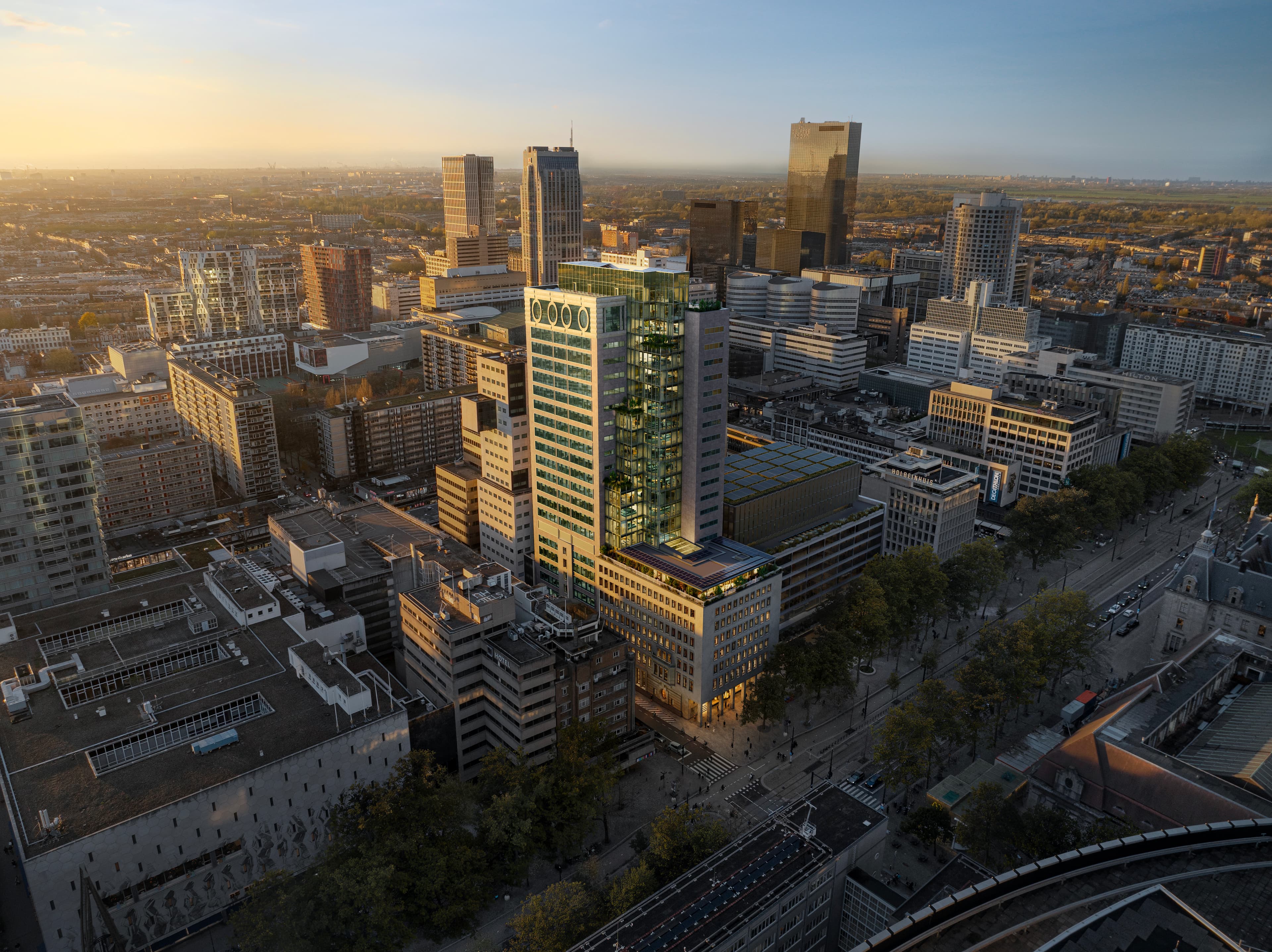 High-angle sunset view of a modern city skyline, featuring a tall glass tower with green terraces centered among mid-rise office blocks and a tree-lined boulevard below.